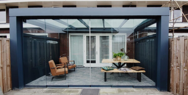 Zadaszenie tarasowe Cube prezentuje eleganckie, przeszklone patio z nowoczesną pergolą, opalonymi fotelami, drewnianą ławką i stołem z roślinami, poduszkami, przesuwanymi drzwiami z białymi żaluzjami i drewnianym ogrodzeniem graniczącym z przestrzenią.