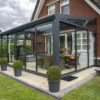 Do ceglanego domu przylega nowoczesny, przeszklony sunroom z meblami ogrodowymi i roślinami doniczkowymi na wyłożonym kafelkami patio z eleganckim Zadaszeniem tarasowym ze szkła, otoczonym zadbanym trawnikiem pod zachmurzonym niebem.