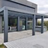 Nowoczesny dom z dużą pergolą-Zadaszenie tarasowe Cube z elektrycznym dachem żaluzjowym na wyłożonym kafelkami patio obok drewnianego tarasu. Szklane drzwi odbijają drzewa, a rośliny doniczkowe i meble ogrodowe są widoczne.
