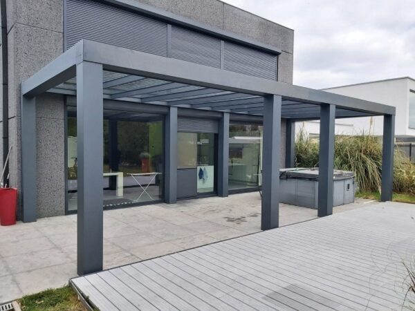 Nowoczesny dom z dużą pergolą-Zadaszenie tarasowe Cube z elektrycznym dachem żaluzjowym na wyłożonym kafelkami patio obok drewnianego tarasu. Szklane drzwi odbijają drzewa, a rośliny doniczkowe i meble ogrodowe są widoczne.