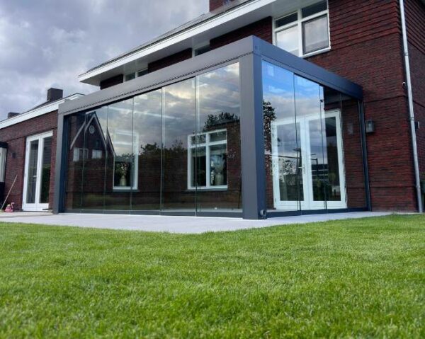 Nowoczesny dom z ceglaną elewacją i dużymi przesuwanymi szklanymi drzwiami posiada przeszklone patio i Zadaszenie tarasowe Cube, otwierające się na świeżo skoszony zielony trawnik pod zachmurzonym niebem.
