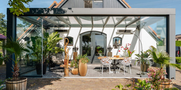 Nowoczesne patio z Zadaszeniem tarasowym Cube, ze szklaną obudową, stołem jadalnym, stylowymi krzesłami, tropikalnymi roślinami i rzeźbami przed białym domem z łukowatymi oknami i drzwiami w słoneczny dzień.