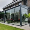 Nowoczesny dom z cegły z przeszklonym patio, na którym znajduje się Zadaszenie tarasowe ze szkła, meble ogrodowe i wiszące krzesło z jajkiem. Przestrzeń podkreśla przystrzyżona trawa, palmy w donicach i sąsiednie domy w tle.