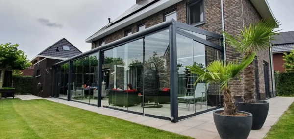 Nowoczesny dom z cegły z przeszklonym patio, na którym znajduje się Zadaszenie tarasowe ze szkła, meble ogrodowe i wiszące krzesło z jajkiem. Przestrzeń podkreśla przystrzyżona trawa, palmy w donicach i sąsiednie domy w tle.
