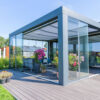 Nowoczesne patio na świeżym powietrzu z przeszkloną obudową i eleganckim Zadaszeniem tarasowym Cube, ozdobione żywymi kwiatami doniczkowymi i wygodnymi siedzeniami, otoczone zielenią i drzewami pod czystym błękitnym niebem.