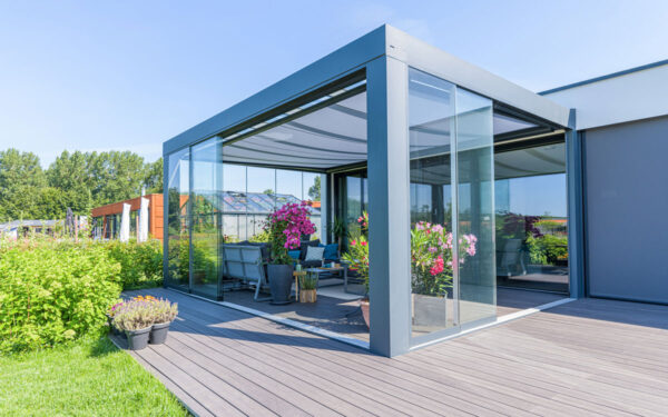 Nowoczesne patio na świeżym powietrzu z przeszkloną obudową i eleganckim Zadaszeniem tarasowym Cube, ozdobione żywymi kwiatami doniczkowymi i wygodnymi siedzeniami, otoczone zielenią i drzewami pod czystym błękitnym niebem.